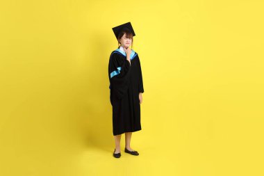 Sarı arka planda hastalıklı bir jestle mavi mezuniyet elbisesi ve kepleri olan bir üniversite öğrencisi. Gen Z, yeni mezun, eğitim, ilk iş