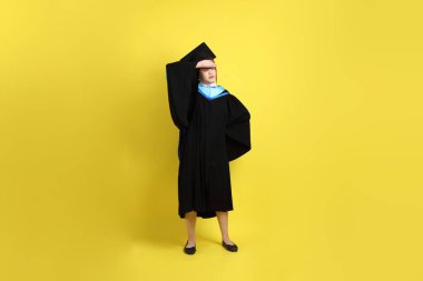 Sarı arka planda hastalıklı bir jestle mavi mezuniyet elbisesi ve kepleri olan bir üniversite öğrencisi. Gen Z, yeni mezun, eğitim, ilk iş