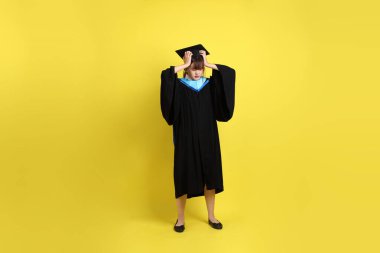 Sarı arka planda hastalıklı bir jestle mavi mezuniyet elbisesi ve kepleri olan bir üniversite öğrencisi. Gen Z, yeni mezun, eğitim, ilk iş