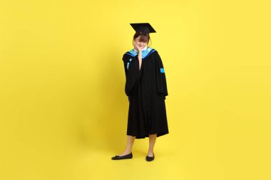 Sarı arka planda diş ağrısı jesti olan mavi mezuniyet elbisesi ve şapkalı bir üniversite öğrencisi. Gen Z, yeni mezun, eğitim, ilk iş