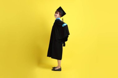 Sarı arka planda sırtı ağrıyan mavi önlüklü ve kepli bir üniversite öğrencisi. Gen Z, yeni mezun, eğitim, ilk iş