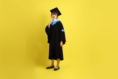 Mavi mezuniyet elbisesi ve kepleri içinde, sarı arka planda sıcak bir izolasyon havası taşıyan bir üniversite öğrencisi. Gen Z, yeni mezun, eğitim, ilk iş