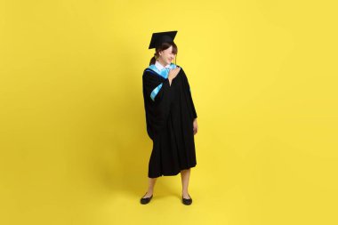 Mavi mezuniyet elbisesi ve kepleri içinde göğüs ağrısı jesti ile sarı arka planda izole edilmiş bir üniversite öğrencisi. Gen Z, yeni mezun, eğitim, ilk iş