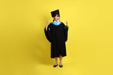 Mavi mezuniyet elbisesi ve kepleri içinde, sarı arka planda güçlü, sağlıklı bir jestle izole edilmiş bir üniversite öğrencisi. Gen Z, yeni mezun, eğitim, ilk iş