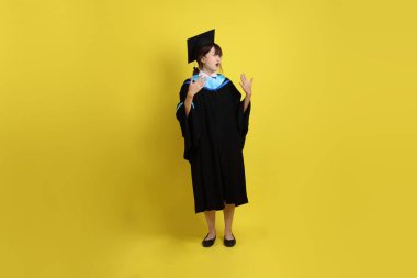 Mavi mezuniyet elbisesi ve kepleri içinde, sarı arka planda sıcak bir izolasyon havası taşıyan bir üniversite öğrencisi. Gen Z, yeni mezun, eğitim, ilk iş