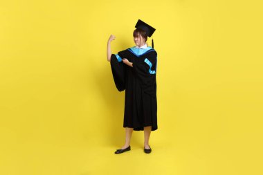 Mavi mezuniyet elbisesi ve kepleri içinde, sarı arka planda güçlü, sağlıklı bir jestle izole edilmiş bir üniversite öğrencisi. Gen Z, yeni mezun, eğitim, ilk iş