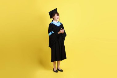 Mavi mezuniyet elbisesi ve şapkalı, sarı arka planda el çırpma hareketi yapan bir üniversite öğrencisi. Gen Z, yeni mezun, eğitim, ilk iş