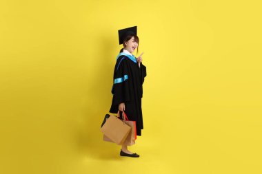 Sarı arka planda, mavi mezuniyet elbiseleri ve kepleri olan, alışveriş çantası taşıyan bir üniversite öğrencisi. Gen Z, yeni mezun, eğitim, ilk iş