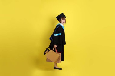 Sarı arka planda, mavi mezuniyet elbiseleri ve kepleri olan, alışveriş çantası taşıyan bir üniversite öğrencisi. Gen Z, yeni mezun, eğitim, ilk iş