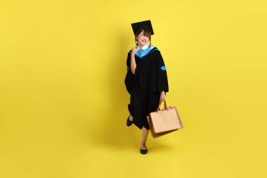 Sarı arka planda, mavi mezuniyet elbiseleri ve kepleri olan, alışveriş çantası taşıyan bir üniversite öğrencisi. Gen Z, yeni mezun, eğitim, ilk iş