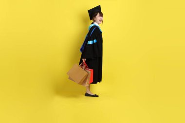 Sarı arka planda, mavi mezuniyet elbiseleri ve kepleri olan, alışveriş çantası taşıyan bir üniversite öğrencisi. Gen Z, yeni mezun, eğitim, ilk iş