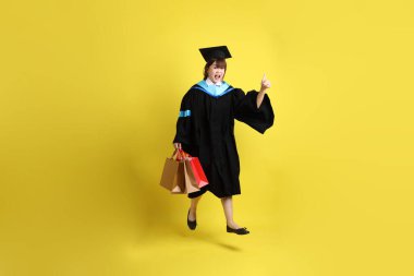 Sarı arka planda, mavi mezuniyet elbiseleri ve kepleri olan, alışveriş çantası taşıyan bir üniversite öğrencisi. Gen Z, yeni mezun, eğitim, ilk iş