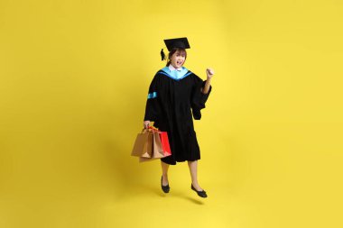 Sarı arka planda, mavi mezuniyet elbiseleri ve kepleri olan, alışveriş çantası taşıyan bir üniversite öğrencisi. Gen Z, yeni mezun, eğitim, ilk iş