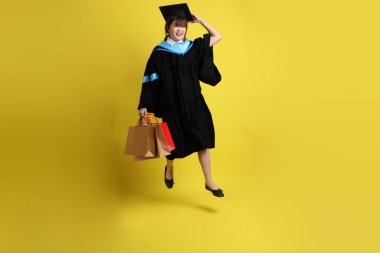 Sarı arka planda, mavi mezuniyet elbiseleri ve kepleri olan, alışveriş çantası taşıyan bir üniversite öğrencisi. Gen Z, yeni mezun, eğitim, ilk iş