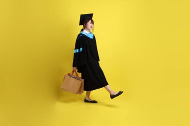 Sarı arka planda, mavi mezuniyet elbiseleri ve kepleri olan, alışveriş çantası taşıyan bir üniversite öğrencisi. Gen Z, yeni mezun, eğitim, ilk iş