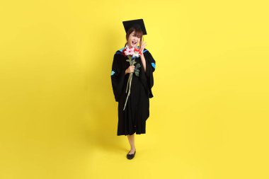 Mavi mezuniyet elbisesi ve kepleri içinde sarı arka planda saklanmış çiçekleri tutan bayan üniversite öğrencisi. Gen Z, yeni mezun, eğitim, ilk iş