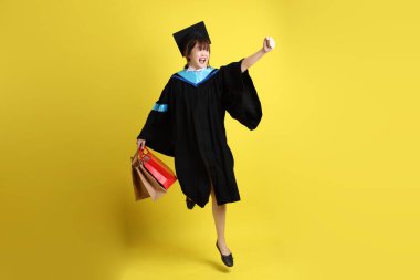 Sarı arka planda, mavi mezuniyet elbiseleri ve kepleri olan, alışveriş çantası taşıyan bir üniversite öğrencisi. Gen Z, yeni mezun, eğitim, ilk iş