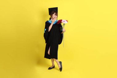 Mavi mezuniyet elbisesi ve kepleri içinde sarı arka planda saklanmış çiçekleri tutan bayan üniversite öğrencisi. Gen Z, yeni mezun, eğitim, ilk iş