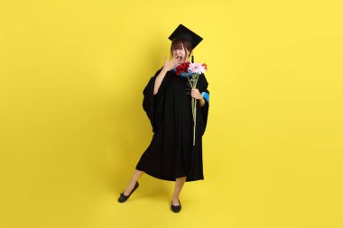 Mavi mezuniyet elbisesi ve kepleri içinde sarı arka planda saklanmış çiçekleri tutan bayan üniversite öğrencisi. Gen Z, yeni mezun, eğitim, ilk iş