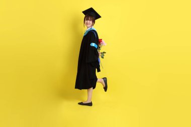 Mavi mezuniyet elbisesi ve kepleri içinde sarı arka planda saklanmış çiçekleri tutan bayan üniversite öğrencisi. Gen Z, yeni mezun, eğitim, ilk iş