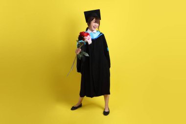 Mavi mezuniyet elbisesi ve kepleri içinde sarı arka planda saklanmış çiçekleri tutan bayan üniversite öğrencisi. Gen Z, yeni mezun, eğitim, ilk iş