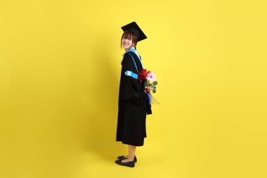 Mavi mezuniyet elbisesi ve kepleri içinde sarı arka planda saklanmış çiçekleri tutan bayan üniversite öğrencisi. Gen Z, yeni mezun, eğitim, ilk iş