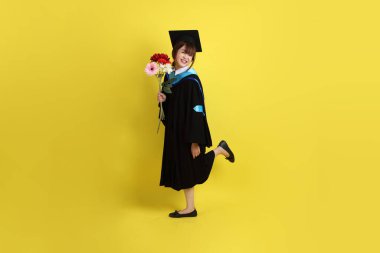 Mavi mezuniyet elbisesi ve kepleri içinde sarı arka planda saklanmış çiçekleri tutan bayan üniversite öğrencisi. Gen Z, yeni mezun, eğitim, ilk iş