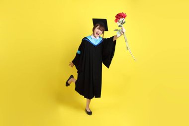 Mavi mezuniyet elbisesi ve kepleri içinde sarı arka planda saklanmış çiçekleri tutan bayan üniversite öğrencisi. Gen Z, yeni mezun, eğitim, ilk iş