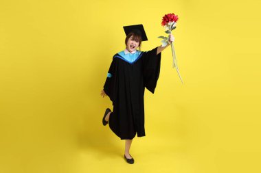 Mavi mezuniyet elbisesi ve kepleri içinde sarı arka planda saklanmış çiçekleri tutan bayan üniversite öğrencisi. Gen Z, yeni mezun, eğitim, ilk iş