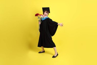 Mavi mezuniyet elbisesi ve kepleri içinde sarı arka planda saklanmış çiçekleri tutan bayan üniversite öğrencisi. Gen Z, yeni mezun, eğitim, ilk iş