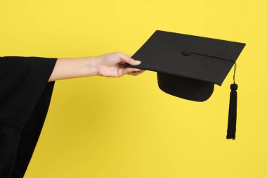 Mavi mezuniyet elbiseli bayan üniversite öğrencisi sarı arka planda izole edilmiş mezuniyet şapkalarının jesti. Gen Z, yeni mezun, eğitim, ilk iş