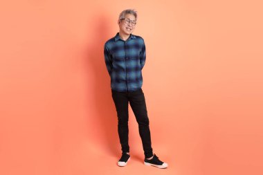 Yeşil ekose giysili Asyalı son sınıf öğrencisi gözlüklü, ayakta duran ve turuncu arka planda sırıtan. Turist, akıllı gündelik.