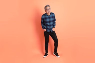 Yeşil ekose giysili Asyalı son sınıf öğrencisi gözlüklü, ayakta duran ve turuncu arka planda sırıtan. Turist, akıllı gündelik.