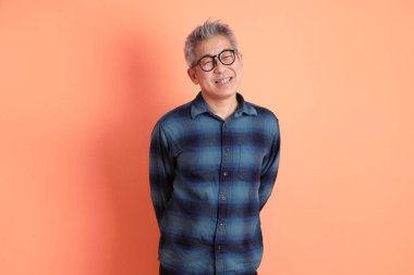 Yeşil ekose giysili Asyalı son sınıf öğrencisi gözlüklü, ayakta duran ve turuncu arka planda sırıtan. Turist, akıllı gündelik.