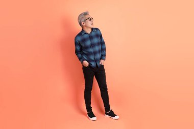 Yeşil ekose giysili Asyalı son sınıf öğrencisi gözlüklü, ayakta duran ve turuncu arka planda sırıtan. Turist, akıllı gündelik.