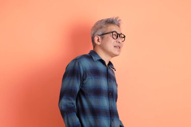 Yeşil ekose giysili Asyalı son sınıf öğrencisi gözlüklü, ayakta duran ve turuncu arka planda sırıtan. Turist, akıllı gündelik.