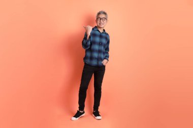 Yeşil ekose elbiseli, gözlüklü, turuncu arka planda izole edilmiş bir yere işaret eden Asyalı son sınıf öğrencisi. Turist, akıllı gündelik.