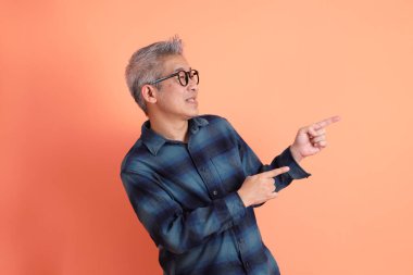 Yeşil ekose elbiseli, gözlüklü, turuncu arka planda izole edilmiş bir yere işaret eden Asyalı son sınıf öğrencisi. Turist, akıllı gündelik.