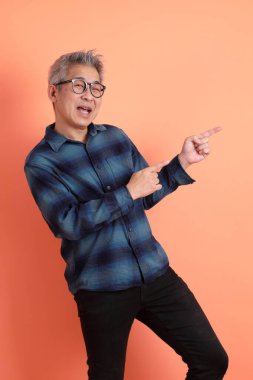 Yeşil ekose elbiseli, gözlüklü, turuncu arka planda izole edilmiş bir yere işaret eden Asyalı son sınıf öğrencisi. Turist, akıllı gündelik.