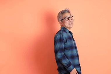 Yeşil ekose giysili Asyalı son sınıf öğrencisi gözlüklü, ayakta duran ve turuncu arka planda sırıtan. Turist, akıllı gündelik.