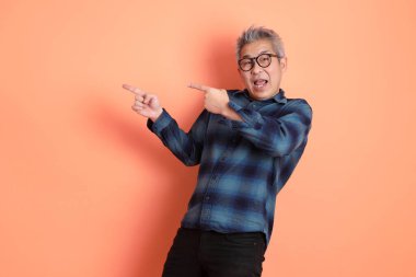 Yeşil ekose elbiseli, gözlüklü, turuncu arka planda izole edilmiş bir yere işaret eden Asyalı son sınıf öğrencisi. Turist, akıllı gündelik.