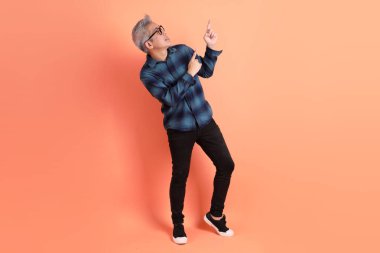 Yeşil ekose elbiseli, gözlüklü, turuncu arka planda izole edilmiş bir yere işaret eden Asyalı son sınıf öğrencisi. Turist, akıllı gündelik.