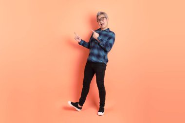 Yeşil ekose elbiseli, gözlüklü, turuncu arka planda izole edilmiş bir yere işaret eden Asyalı son sınıf öğrencisi. Turist, akıllı gündelik.