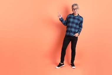 Yeşil ekose elbiseli, gözlüklü, turuncu arka planda izole edilmiş bir yere işaret eden Asyalı son sınıf öğrencisi. Turist, akıllı gündelik.