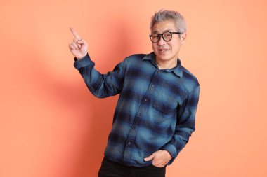 Yeşil ekose elbiseli, gözlüklü, turuncu arka planda izole edilmiş bir yere işaret eden Asyalı son sınıf öğrencisi. Turist, akıllı gündelik.