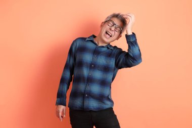 Yeşil ekose giysili Asyalı son sınıf öğrencisi gözlüklü ve turuncu arka planda izole edilmiş stres jesti takıyor. Turist, akıllı gündelik.