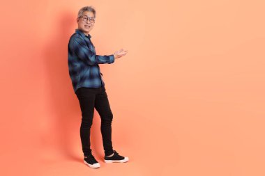 Yeşil ekose elbiseli, gözlüklü, turuncu arka planda izole edilmiş bir yere işaret eden Asyalı son sınıf öğrencisi. Turist, akıllı gündelik.