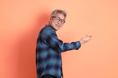 Yeşil ekose elbiseli, gözlüklü, turuncu arka planda izole edilmiş bir yere işaret eden Asyalı son sınıf öğrencisi. Turist, akıllı gündelik.