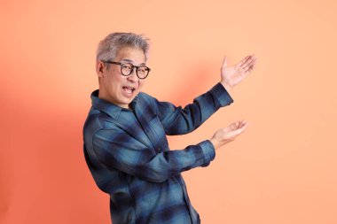 Yeşil ekose elbiseli, gözlüklü, turuncu arka planda izole edilmiş bir yere işaret eden Asyalı son sınıf öğrencisi. Turist, akıllı gündelik.