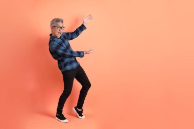 Yeşil ekose elbiseli, gözlüklü, turuncu arka planda izole edilmiş bir yere işaret eden Asyalı son sınıf öğrencisi. Turist, akıllı gündelik.